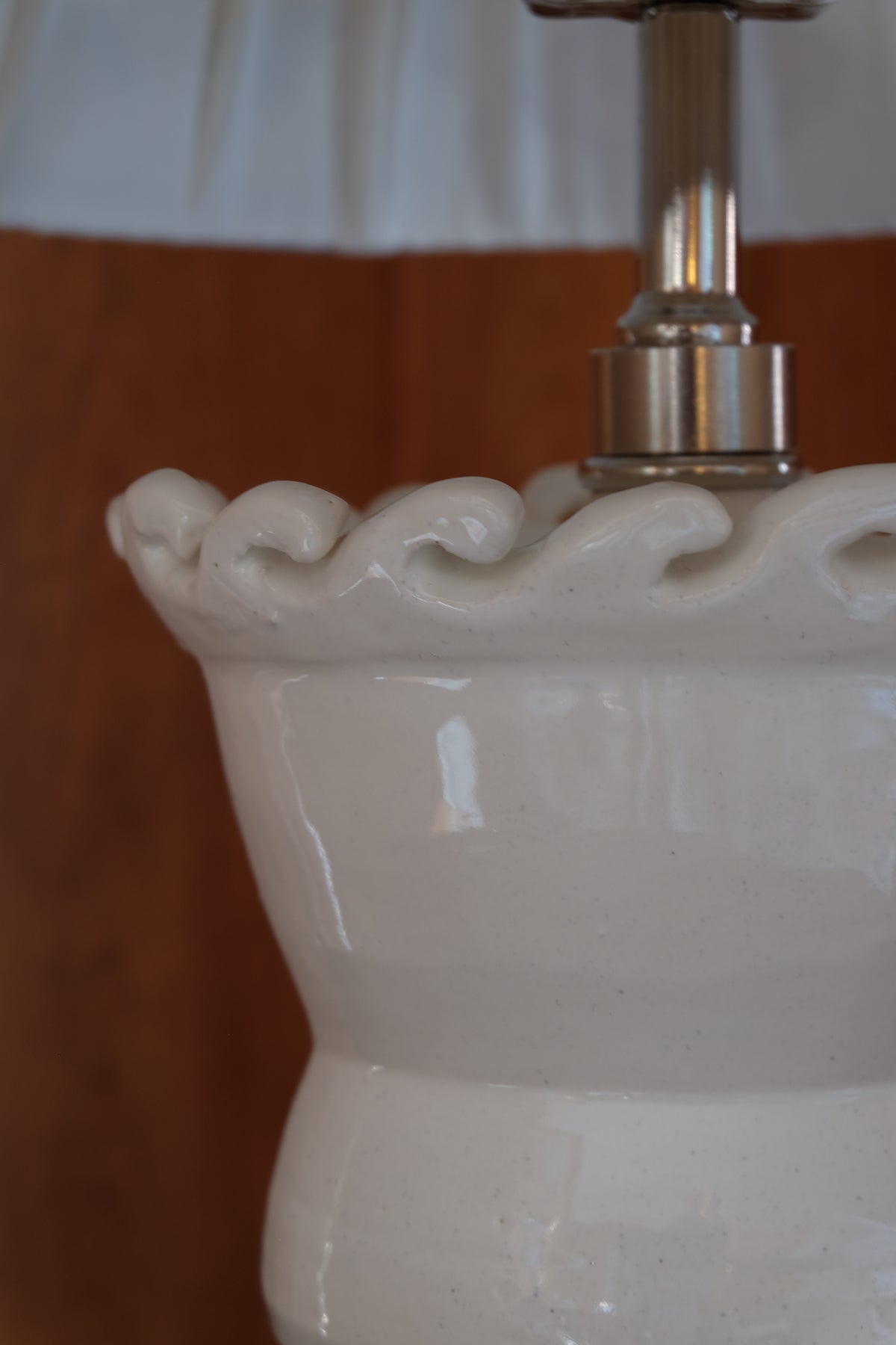 Close-up of a glossy white ceramic lamp base with a decorative wavy rim and a metallic silver-toned socket or hardware piece attached, set against a blurred brown and white background.
